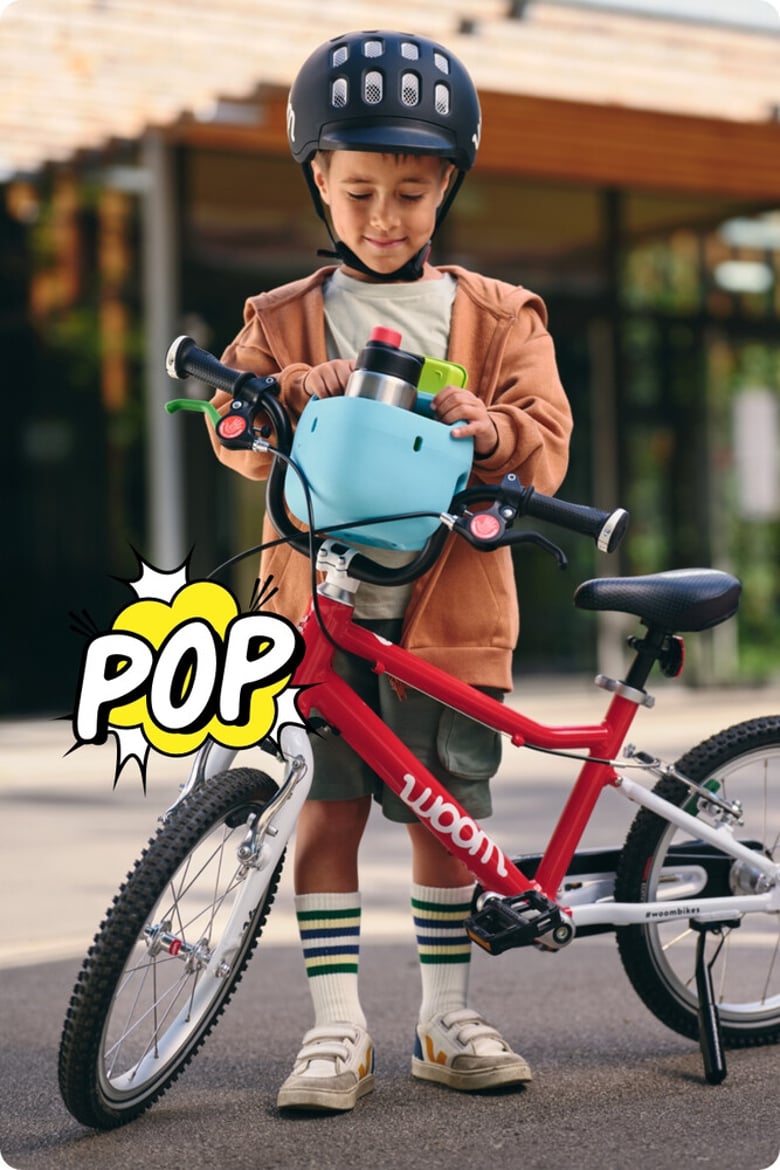 Ein kleiner Junge mit schwarzem Helm steckt einen hellblauen Fahrradkorb mit Trinkflasche auf den Lenker seines rot-weißen woom bikes. Darunter ist eine zerplatzende Comic-Blase mit dem Schriftzug POP.