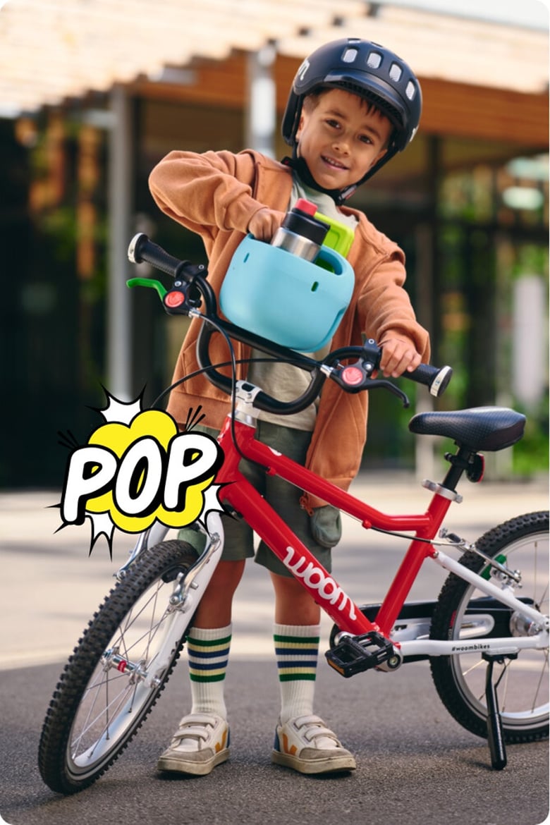 Ein kleiner Junge mit schwarzem Helm steckt einen hellblauen Fahrradkorb mit Trinkflasche vom Lenker seines rot-weißen woom bikes ab. Darunter ist eine zerplatzende Comic-Blase mit dem Schriftzug POP.