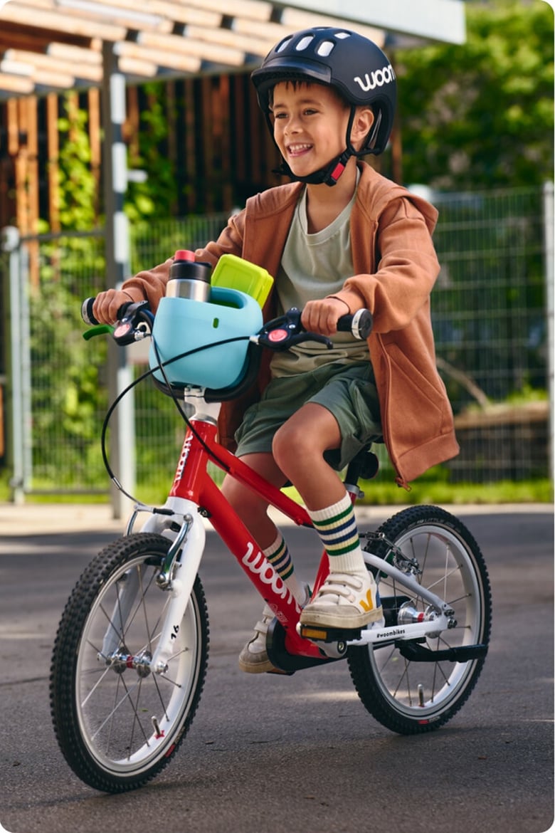 Ein kleiner Junge mit schwarzem Helm fährt mit einem rot-weißen woom bike, auf dessen Lenker ein hellblauer Fahrradkorb mit Trinkflasche angebracht ist.