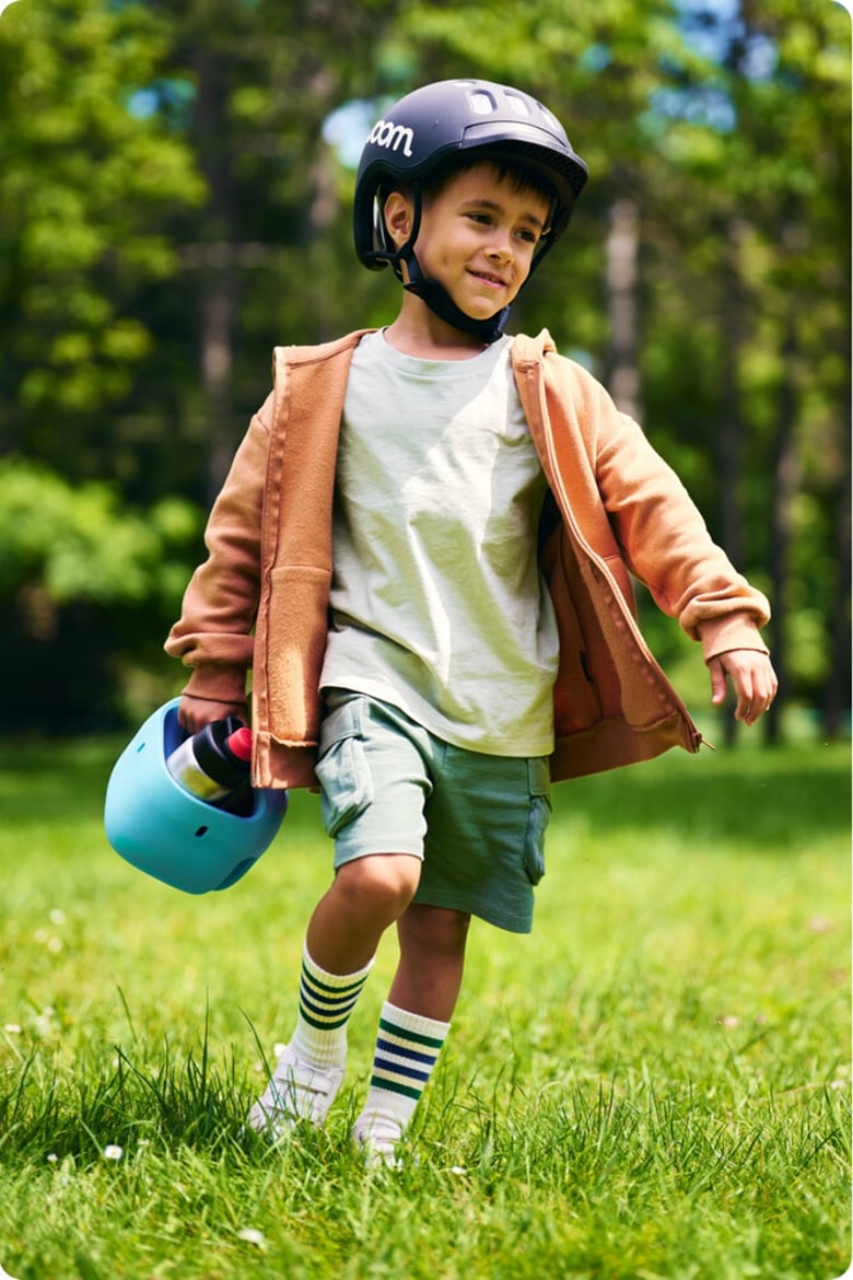 Ein kleiner Junge mit schwarzem Helm läuft über eine Wiese. In der rechten Hand trägt er einen hellblauen Fahrradkorb mit einer Trinkflasche darin.
