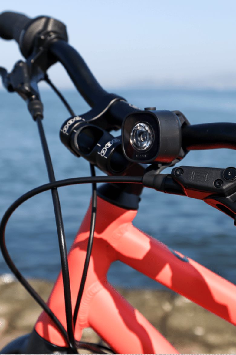 A closeup image of the handlebars, stem, and frame of a neon coral woom EXPLORE bike with a BEAM Bike Light attached, parked in front of the San Francisco Bay.