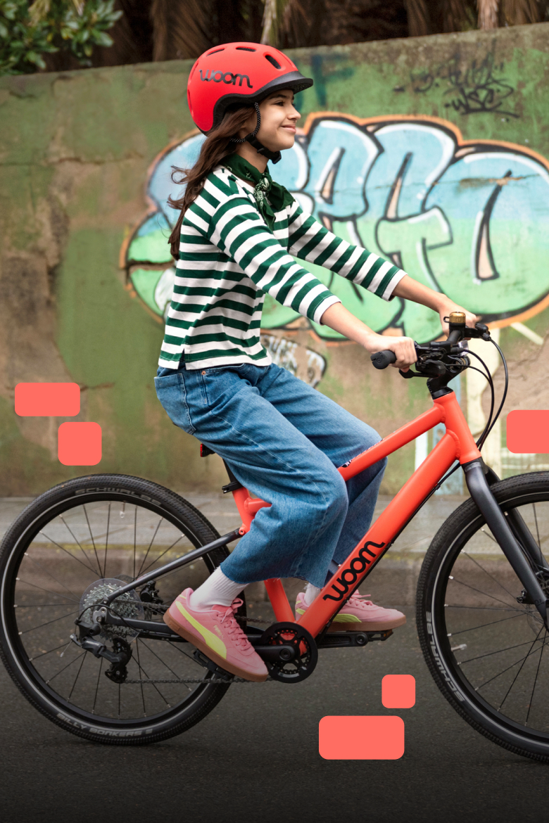 A smiling child wearing a woom READY Helmet rides a coral EXPLORE past a graffiti-covered wall.