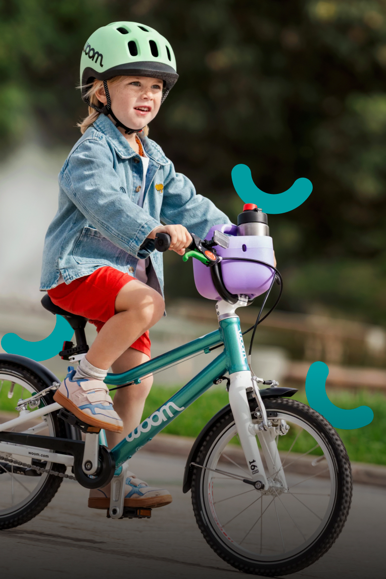 A kid wearing a READY helmet rides a woom GO bicycle with a purple POP basket outdoors.