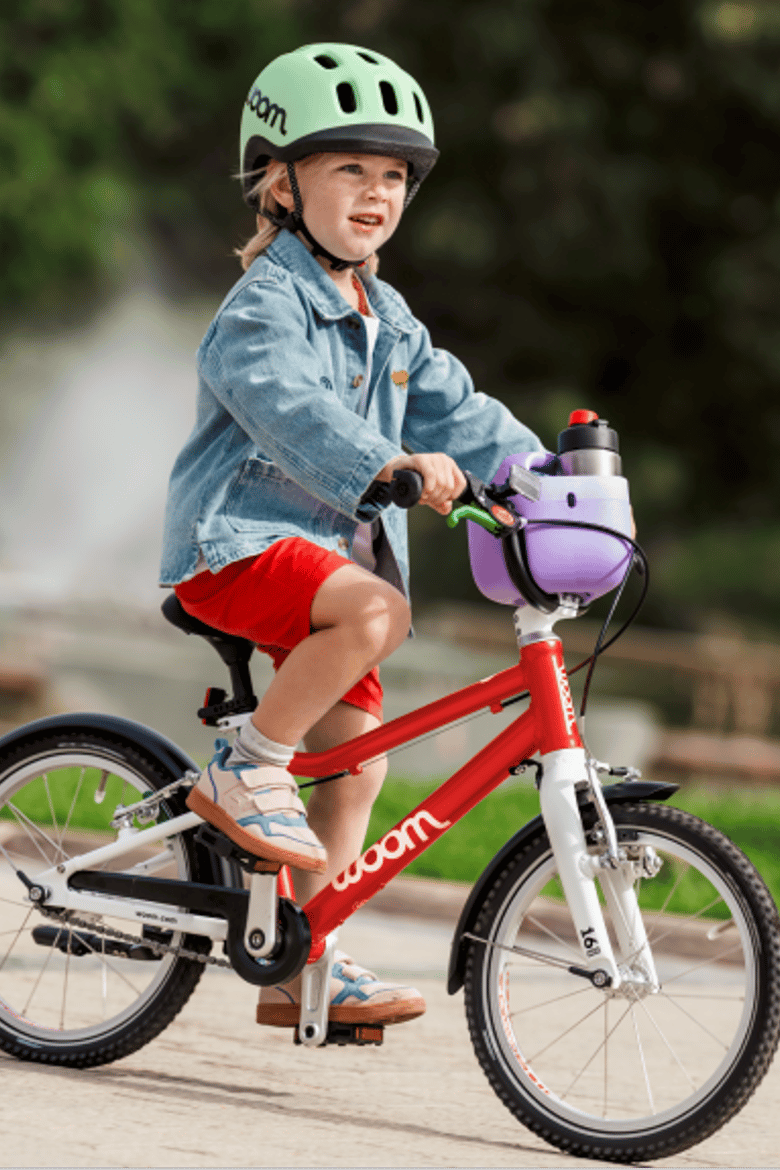 A young child wearing a mint READY Helmet rides a red woom GO 3 past a fountain in a lush green park. The bike is equipped with a wild violet POP Basket and GLUG water bottle. 