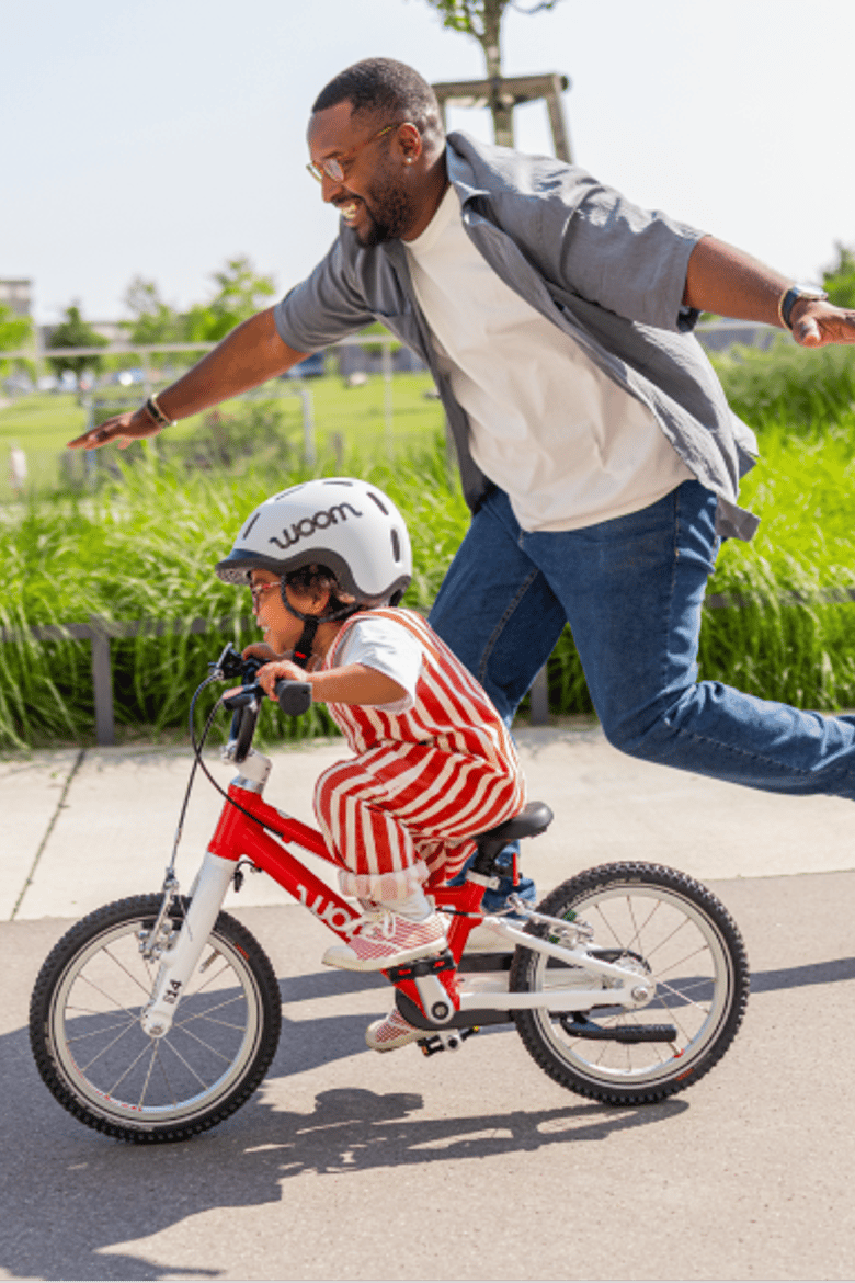 A father runs cheerfully next to his young son as he successfully pedals his red woom GO 2 down an empty street next to a park.