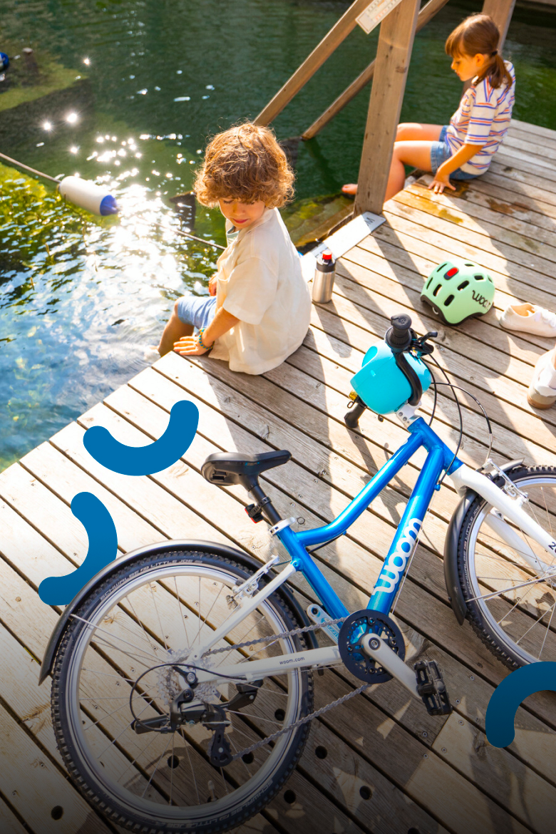 Kids on a pier with a blue woom GO bike and mint woom READY helmet. Sunlight reflects on the sparkling water.
