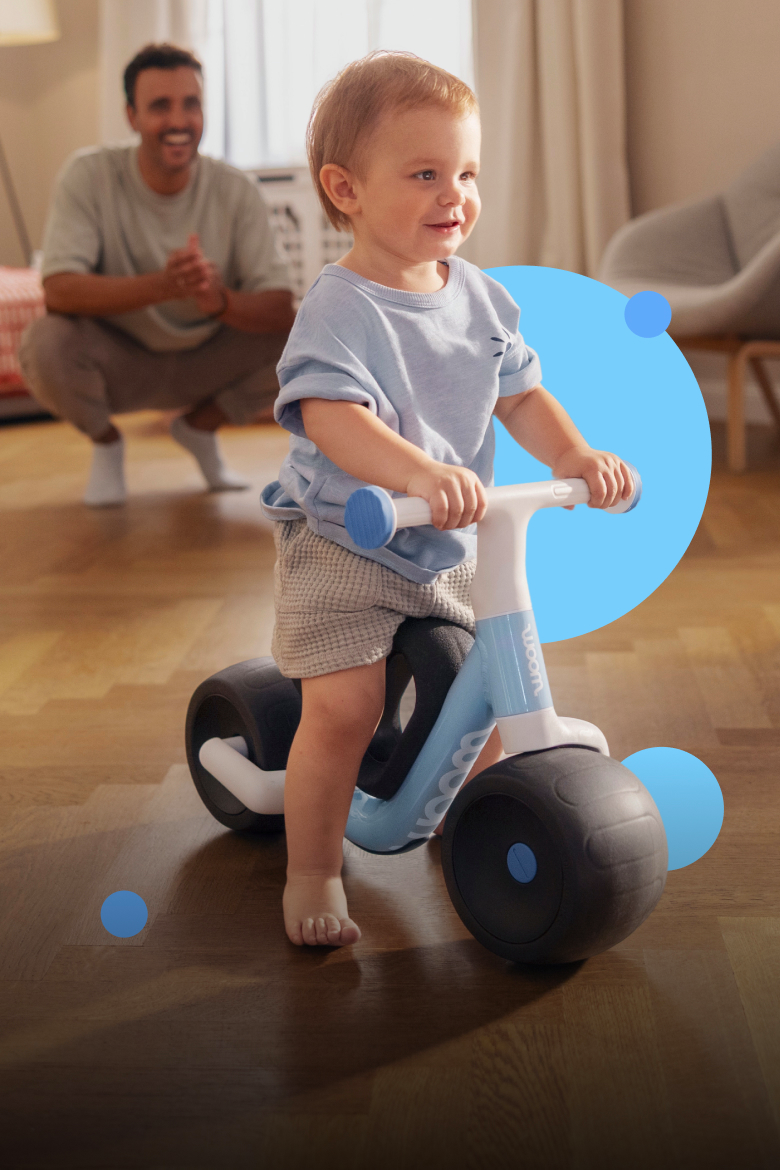 A happy child riding a blue woom WOW in a living room, with an adult watching behind them.