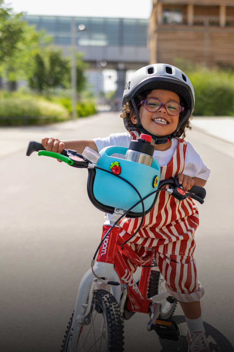 A smiling kid wearing a READY helmet and glasses on a red GO bicycle with a blue POP basket outdoors.
