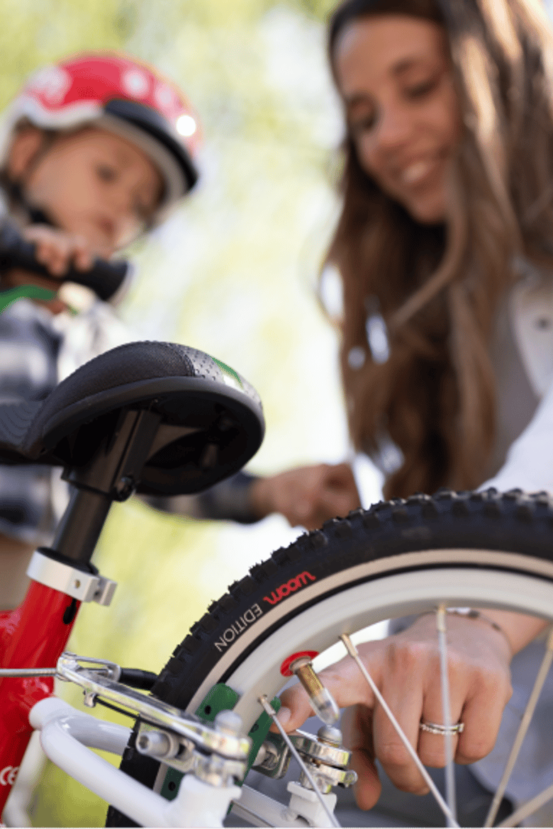 A mom kneels next to a woom bike and touches the brake mechanism while her young son looks on.