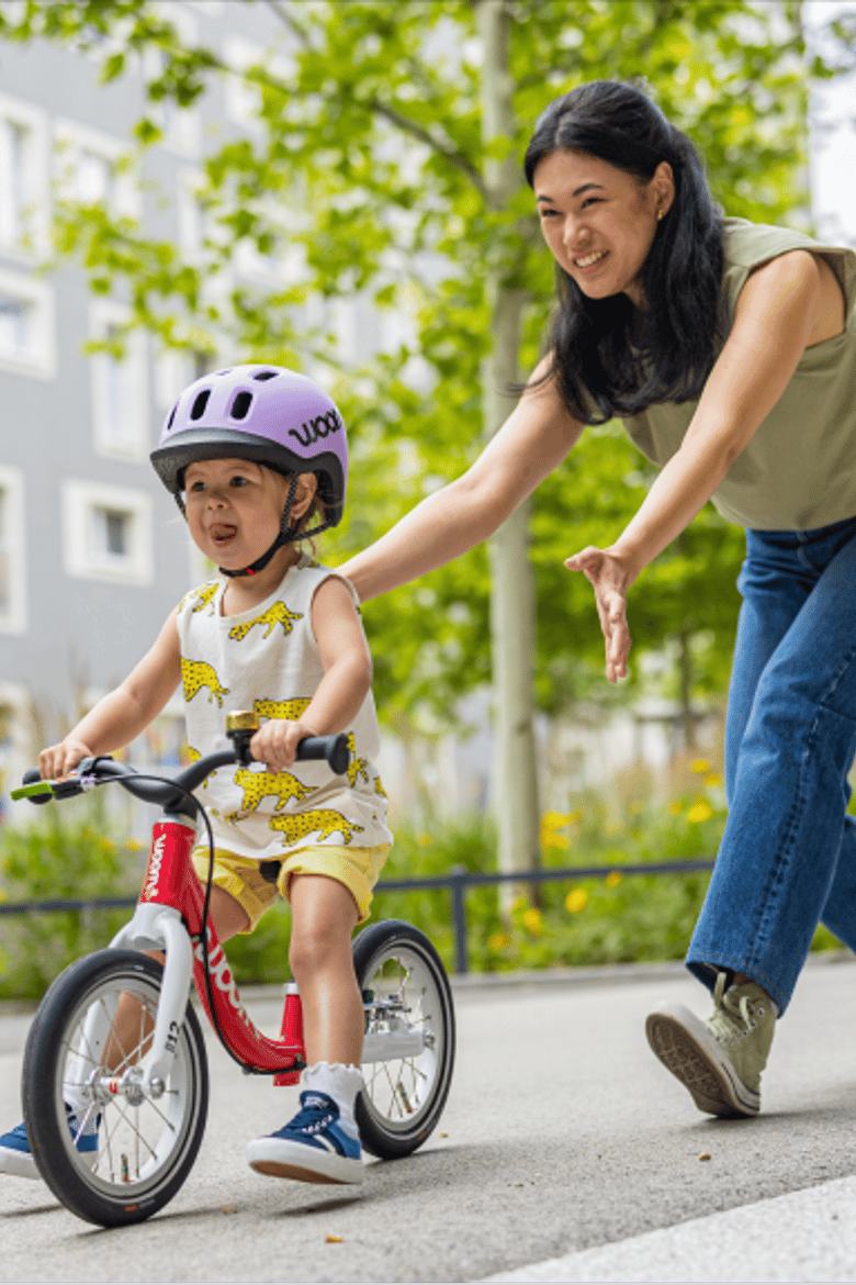 A young girl in a lilac READY Helmet sticks her tongue out in concentration while maneuvering her hot pink woom GO 1 down a sidewalk as her mom follows closely behind.