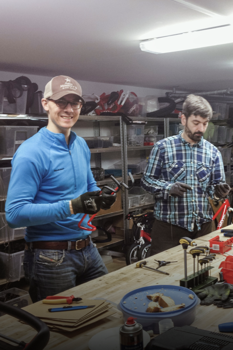 The two woom founders in a bicycle workshop filled with parts, one is smiling holding a red bike accessory.
