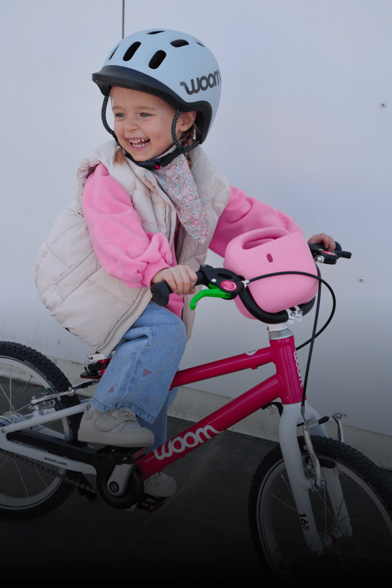 Auf einem rosafarbenen woom GO Kinderfahrrad samt POP Kinderfahrradkorb sitzt ein Mädchen mit grauem READY Helm auf dem Kopf.