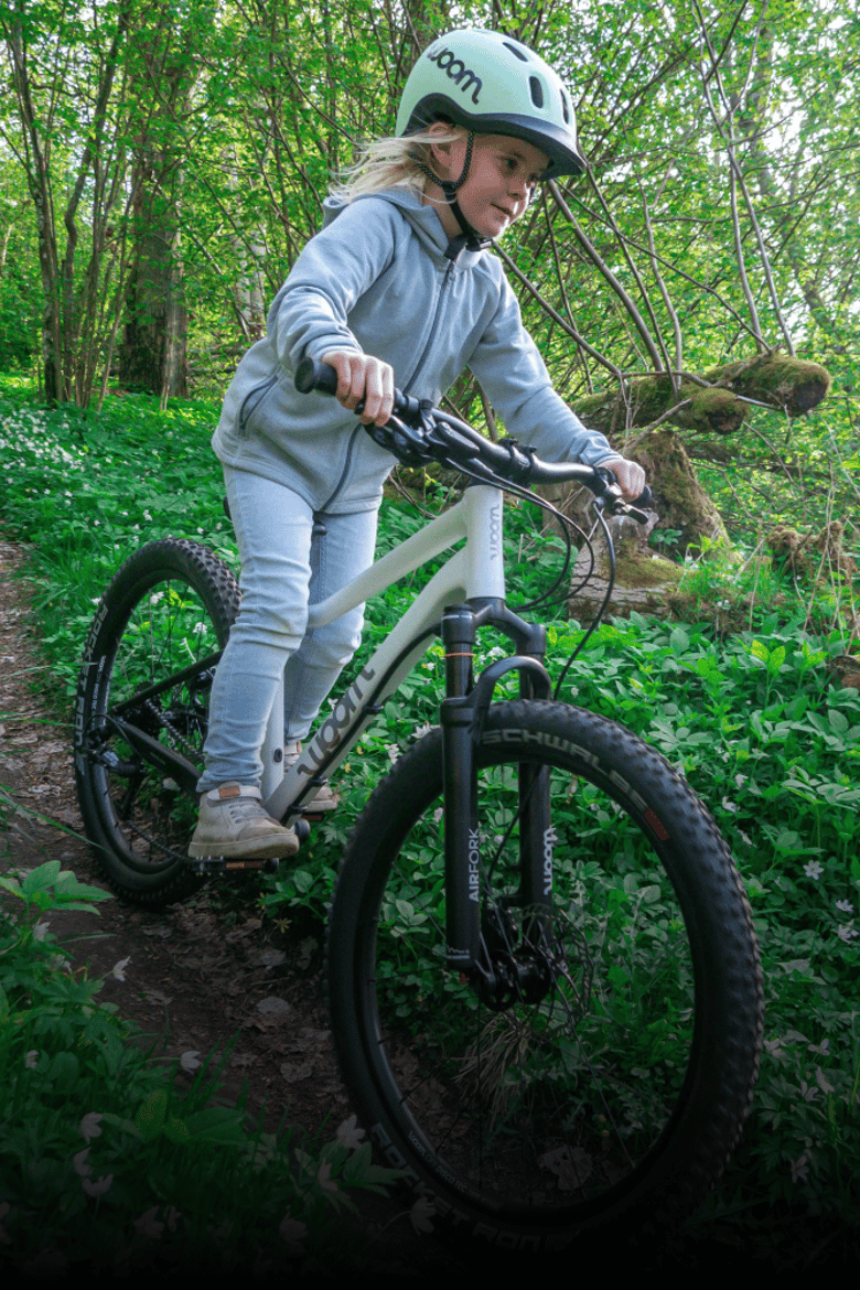 Een meisje met een groene woom READY-kinderhelm rijdt op een woom OFF AIR in de kleur cirrus cloud door het bos.