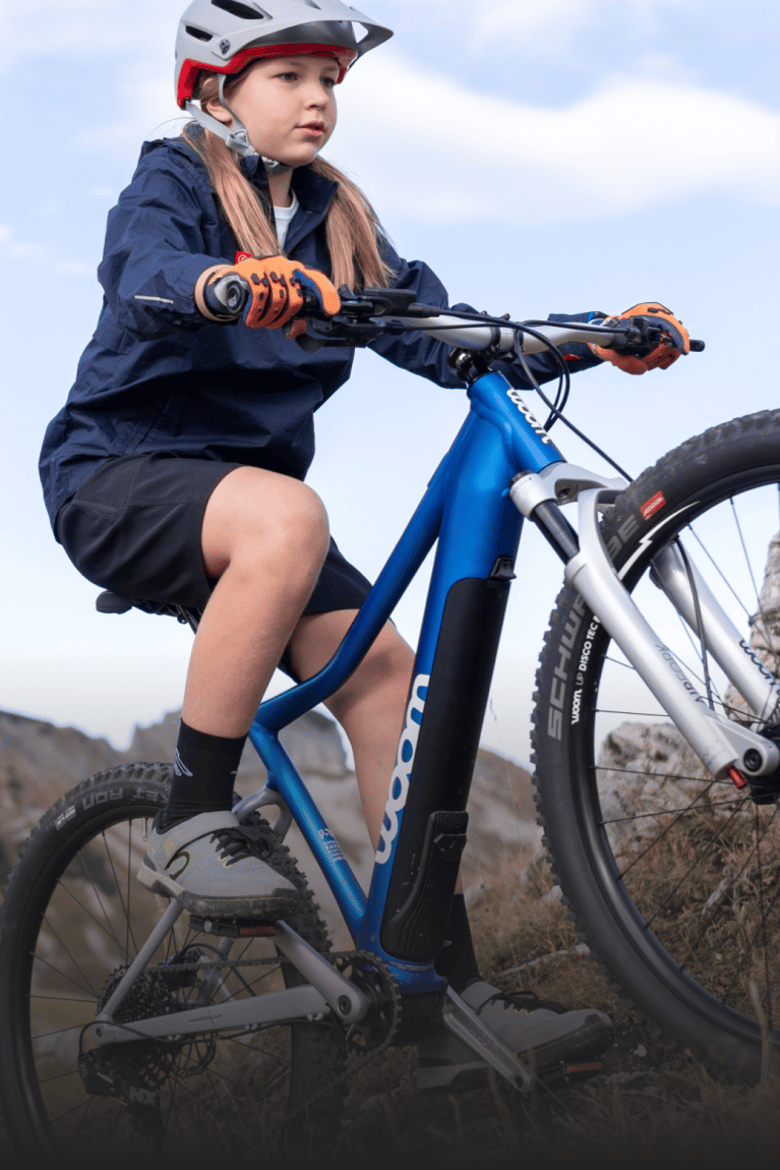Meisje met grijze fietshelm op fietst op een blauwe woom UP E-bike in een bergachtig gebied.