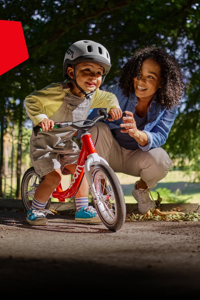 Kleinkind mit grauem READY Kinderhelm auf dem Kopf ist bereit zum Losfahren mit dem woom GO 1, die Mutter lässt es los.