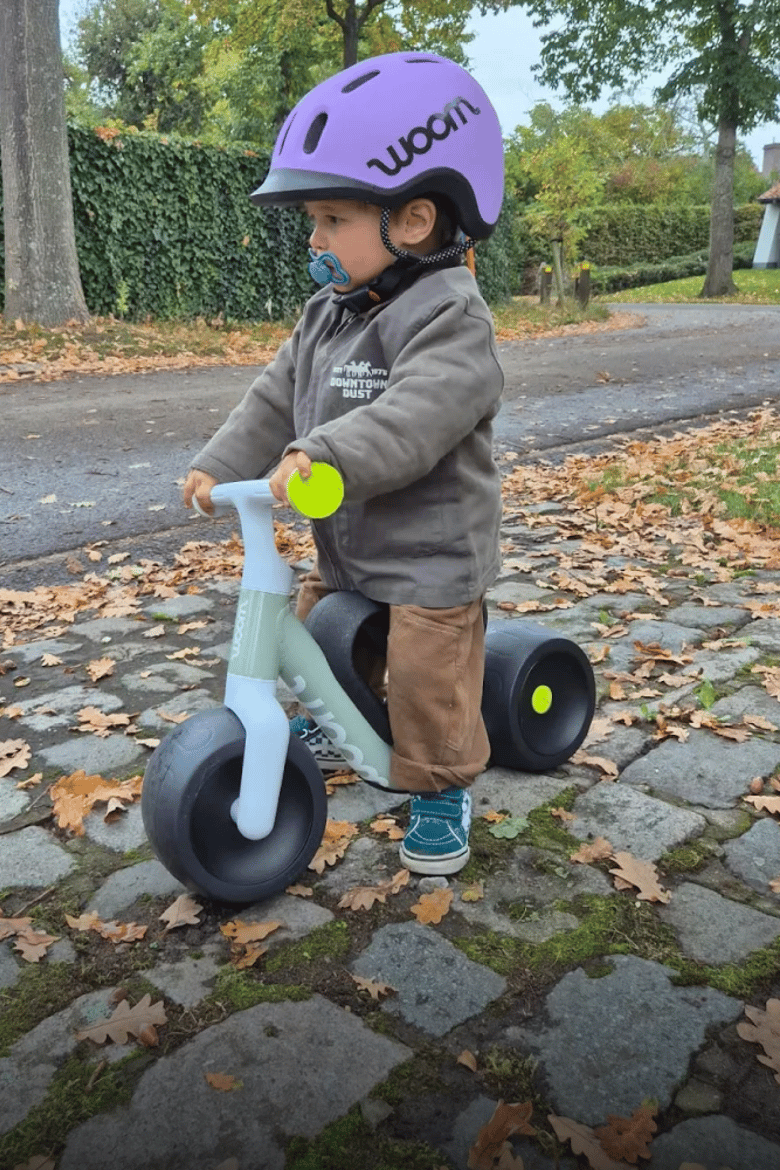 A toddler with a purple READY Kids' Helmet is riding their green woom WOW self-balancing bike on a cobbled pavement.