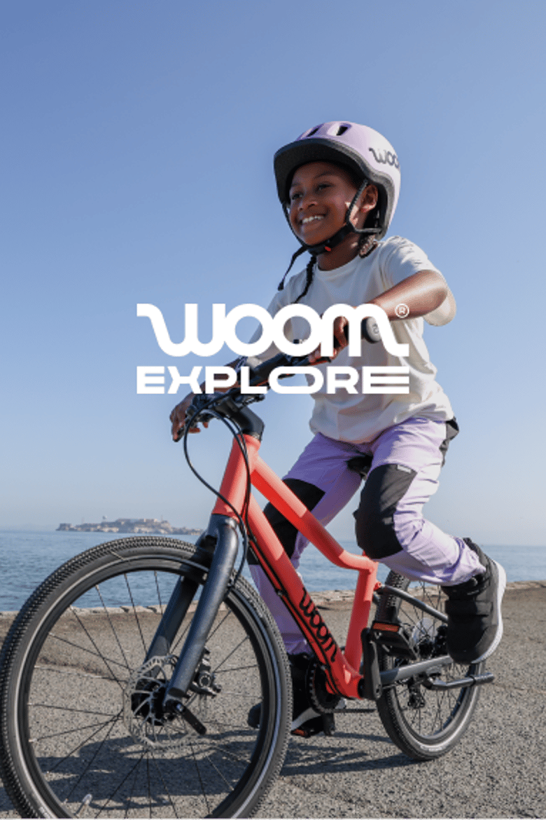 An image of a girl wearing a READY Helmet in lilac while riding her neon coral EXPLORE bike on a paved path next to a serene bay under a bright blue sky.