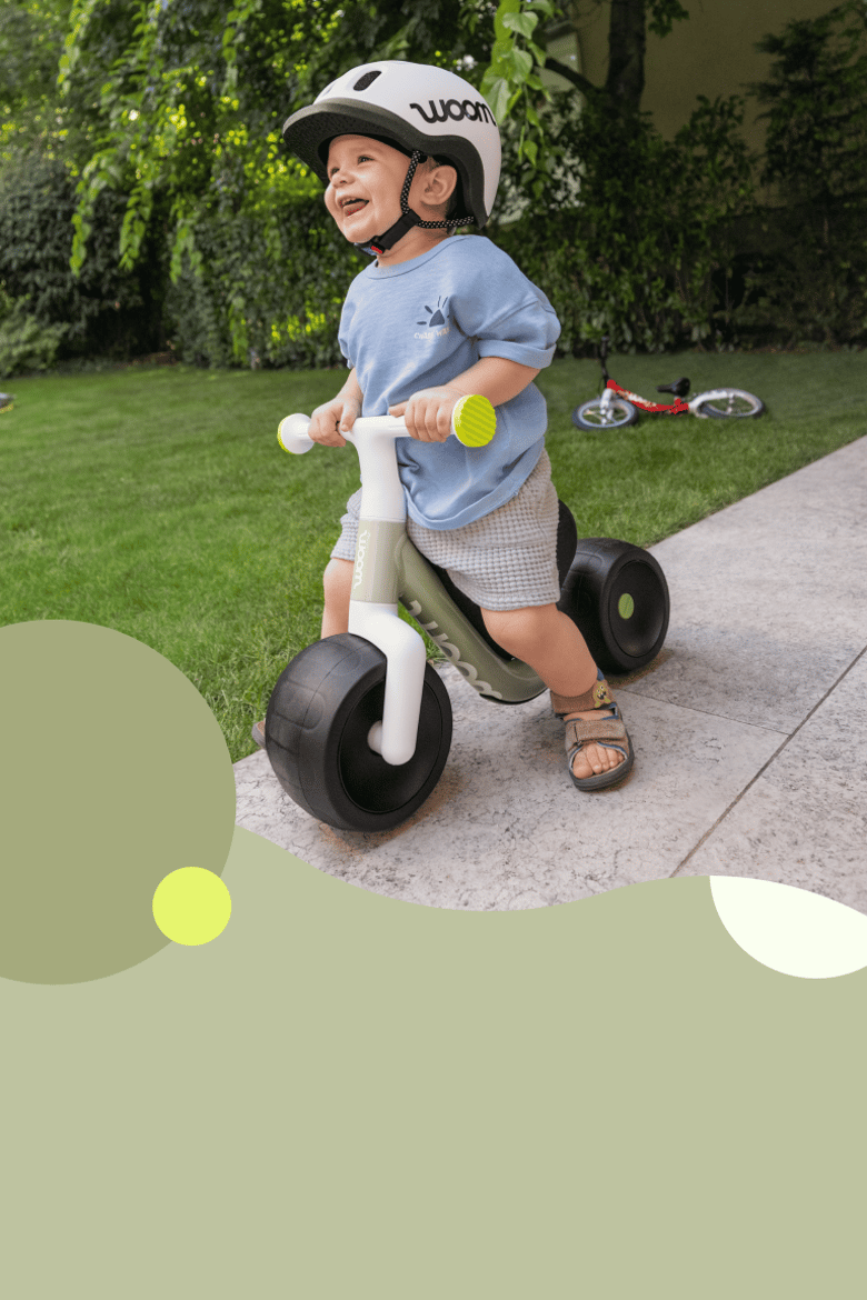 Ett skrattande barn som har barnhjälmen READY på sig kör på terrassen framför trädgården med sin woom WOW i bild.