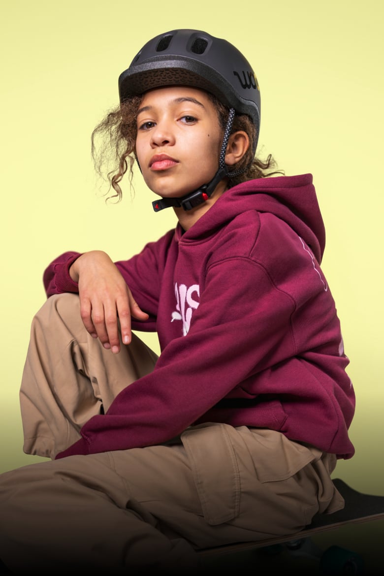 Zittend meisje met zwarte READY-kinderhelm van woom op het hoofd leunt met één arm op een been.