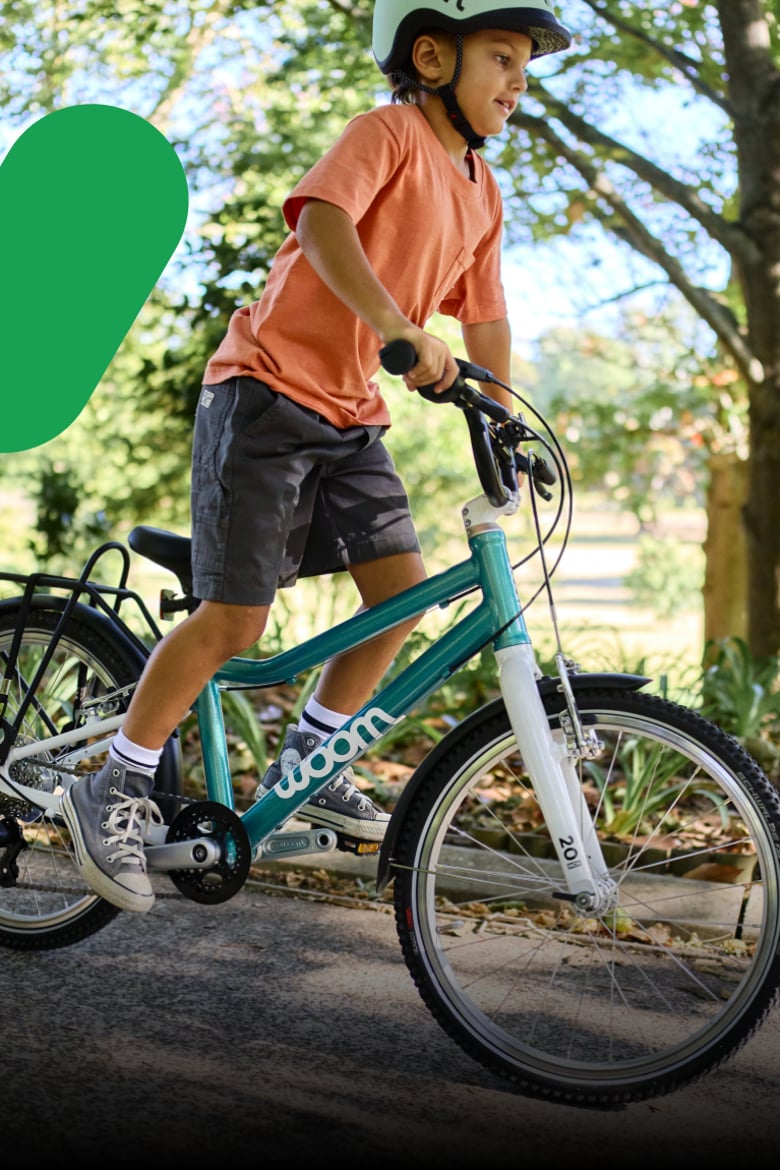 Junge mit grünem READY Kinderhelm auf dem Kopf fährt stehend auf dem woom GO 4 in der Farbe Minzgrün auf einem Weg im Grünen.
