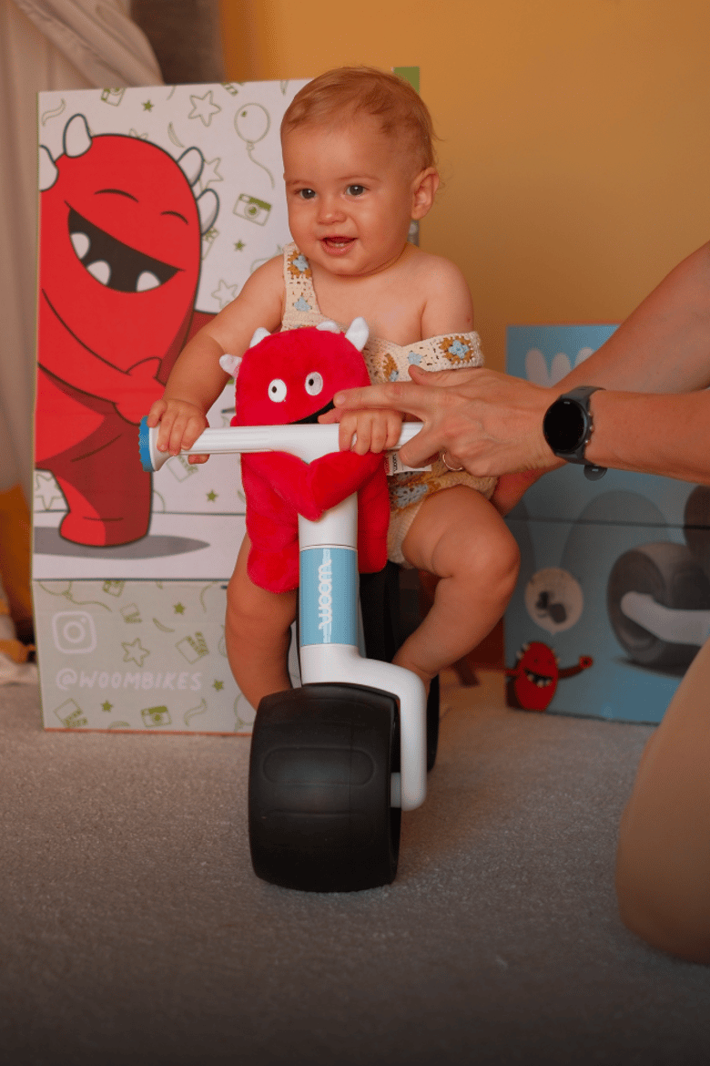 A toddler is sitting on a blue woom WOW with a Buddy softy toy on the handlebars and a Buddy stand in the background.
