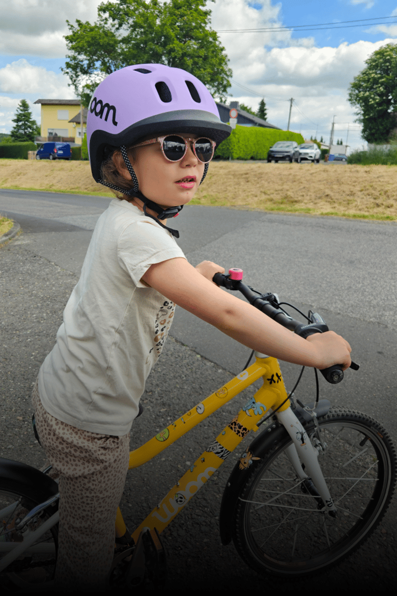 Een kind met een lilakleurige READY-fietshelm op staat bij een gele woom-fiets waarvan het frame met stickers is beplakt.