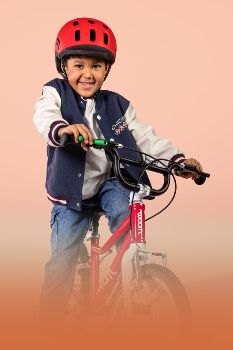 Un jeune garçon souriant et portant un casque READY rouge est assis sur la selle d’un woom GO de couleur rouge.