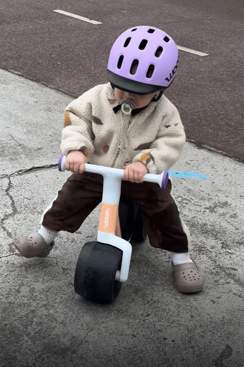 A child with a purple READY Kids' Helmet is riding an orange woom WOW self-balancing bike on the pavement.