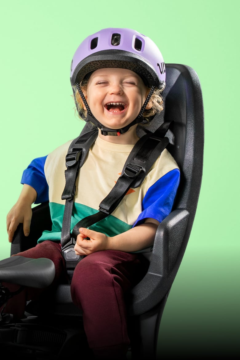 Stralend kind met lila READY-kinderhelm van woom op het hoofd zit in een fietszitje tegen een groene achtergrond.