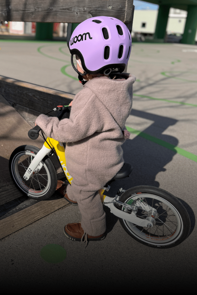 Ein Kleinkind mit lila READY Radhelm und Wollwalkanzug steht auf einem gelben woom GO Laufrad in 12 Zoll.