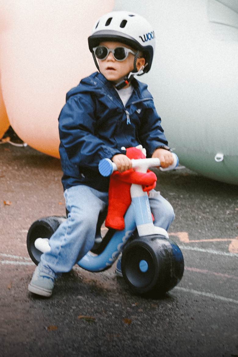 A child with a white READY Kids' Helmet is rolling on a blue woom WOW on tarmac with a red Buddy toy on the handlebars.