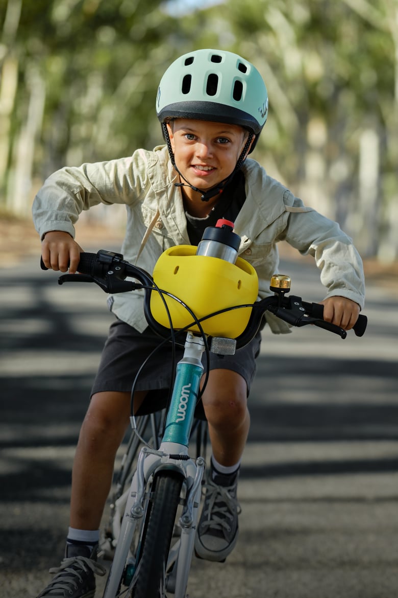 A boy wearing a woom READY helmet, riding a metallic turquoise woom GO bike with vibrant yellow POP basket holding a GLUG water bottle while riding on a road.
