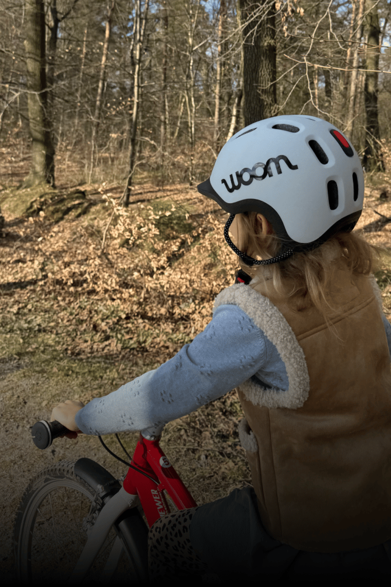 Auf einem roten woom GO Kinderrad fährt ein Kind mit grauem READY Radhelm auf dem Kopf im herbstlichen Wald.