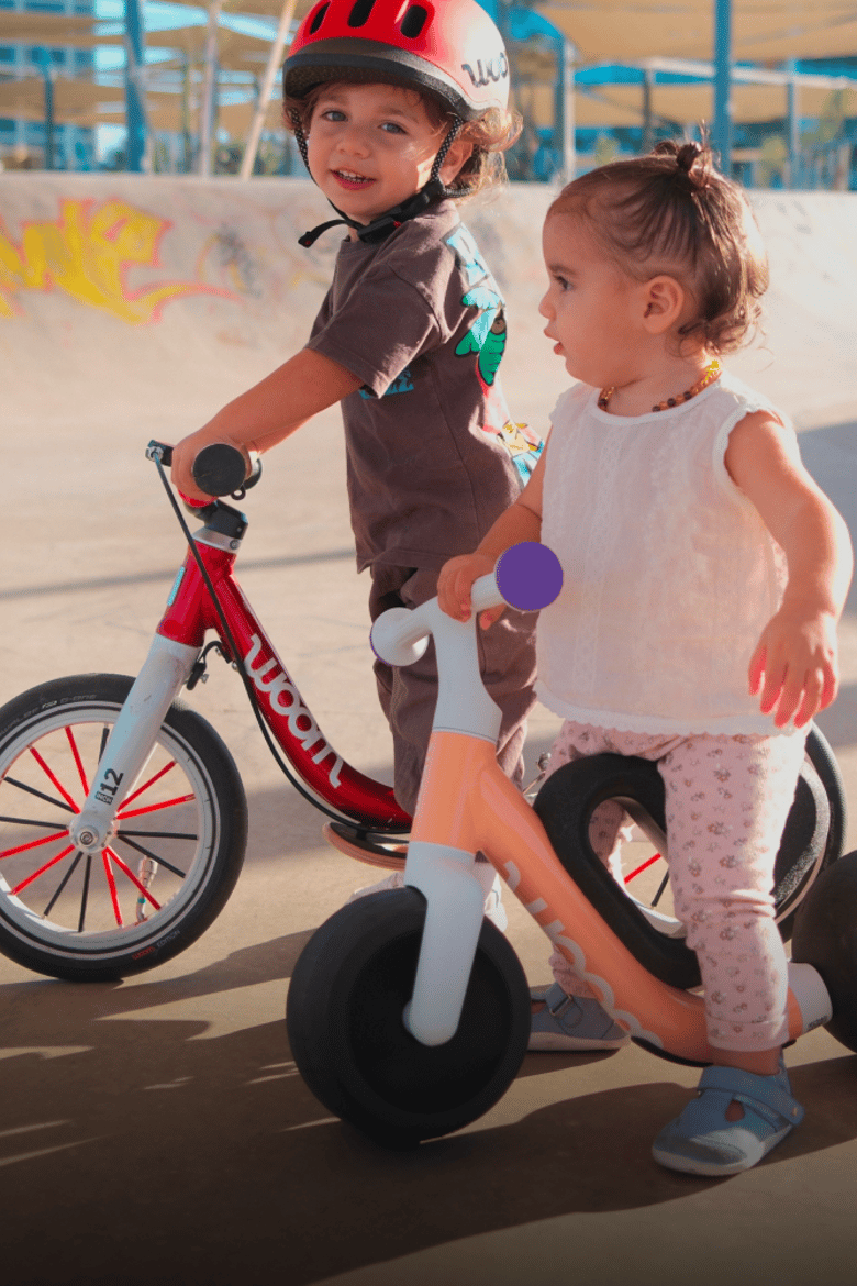 Two toddlers are standing over their balance bikes – one peach woom WOW and one red woom GO 1.
