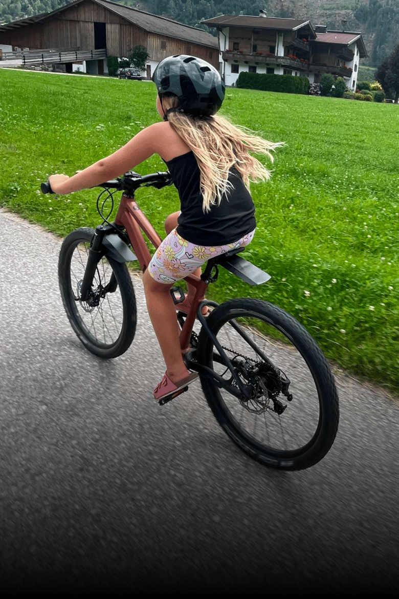 Een meisje met een fietshelm op rijdt op een woom OFF AIR in de kleur terra coppa met MUDBLOCKA-spotborden voor een grasveld.