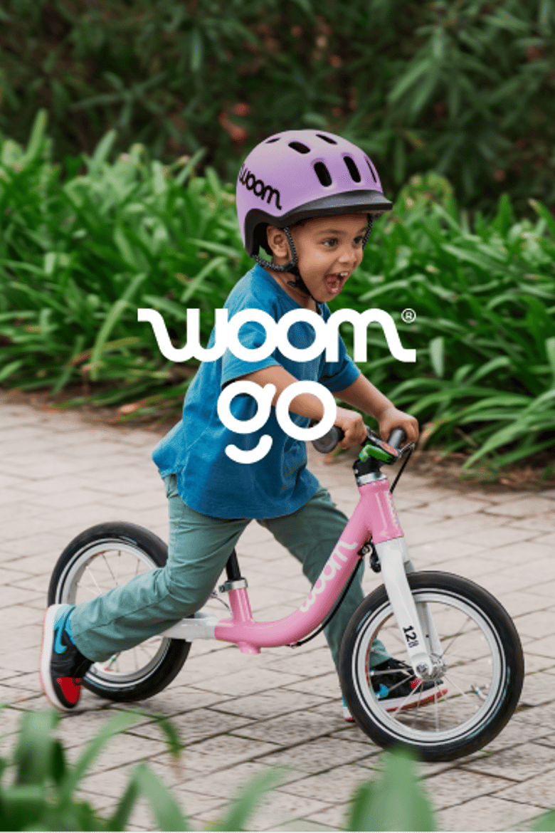 An image of a young boy grinning and wearing a READY Helmet in lilac while riding a powder pink woom GO 1 down a brick path.