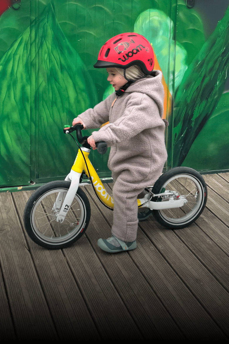 Auf einem gelben woom Laufrad in 12 Zoll rollt ein Kleinkind mit rotem READY Radhelm und Wollwalkanzug.
