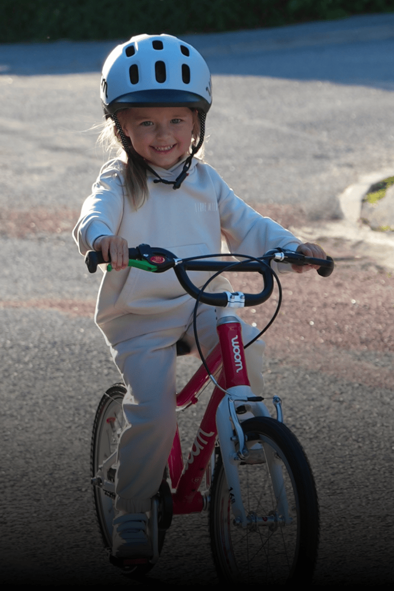 Kind mit grauem READY Radhelm auf dem Kopf fährt mit seinem roten woom GO Fahrrad auf einem Asphaltweg.