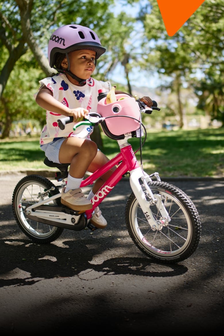 Auf dem Sattel eines pinken woom GO 2 Fahrrads samt Korb sitzt ein Mädchen mit lilafarbenem READY Kinderhelm auf dem Kopf.