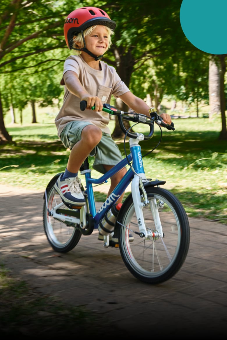 Auf dem Sattel eines blauen woom GO 3 Fahrrads sitzt ein Junge mit rotem READY Kinderhelm auf dem Kopf.
