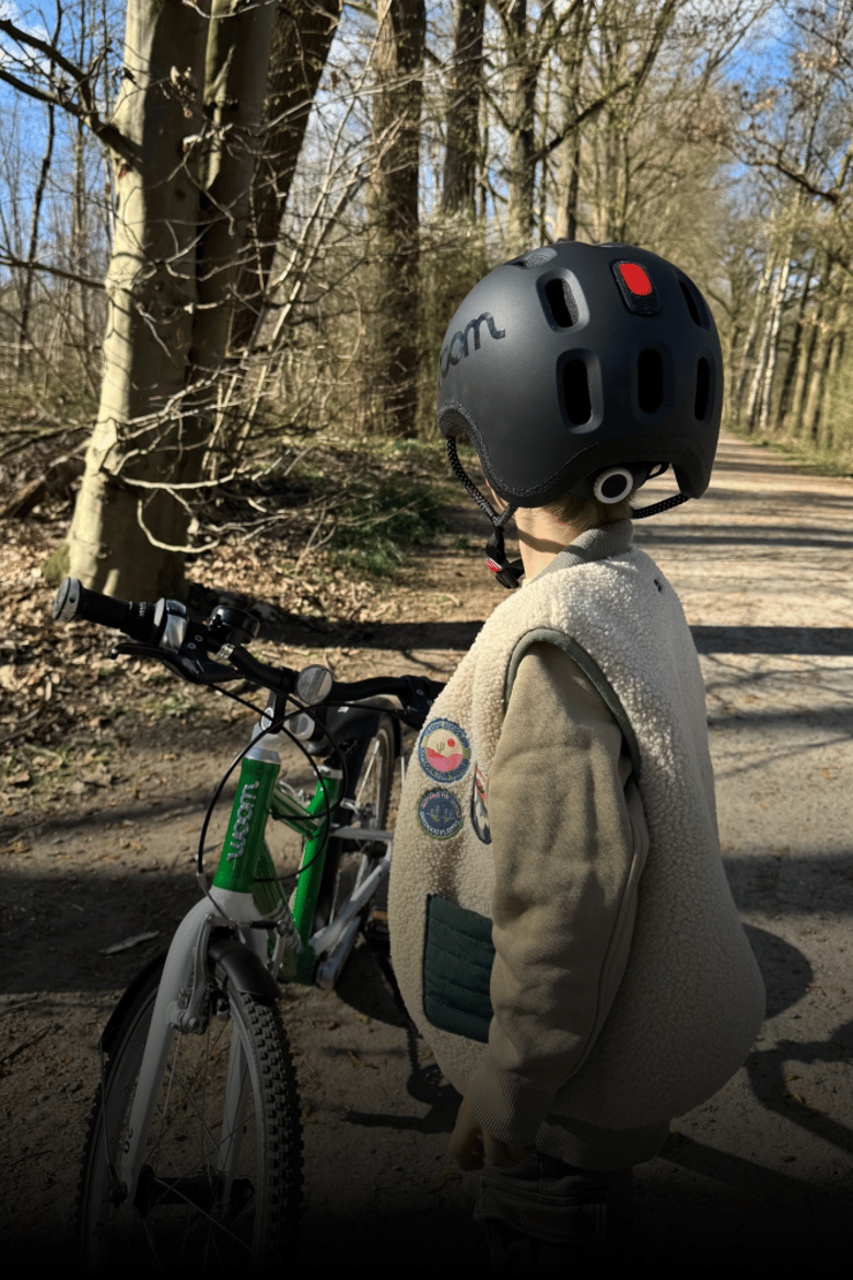 Een kind op de rug gezien met een zwarte READY-helm op met een geïntegreerde helmlamp, en daarachter een groene woom-fiets.