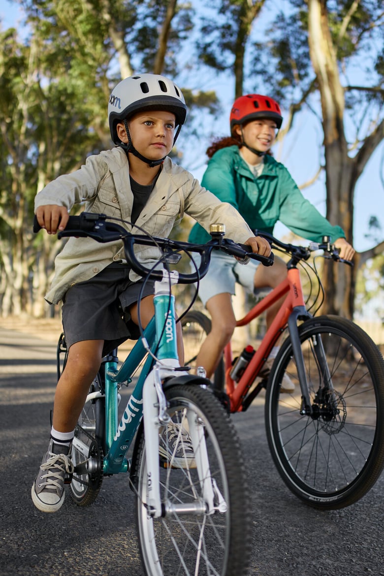 Two children wearing woom READY helmets riding a woom GO and a woom EXPLORE bike along a tree-lined path.