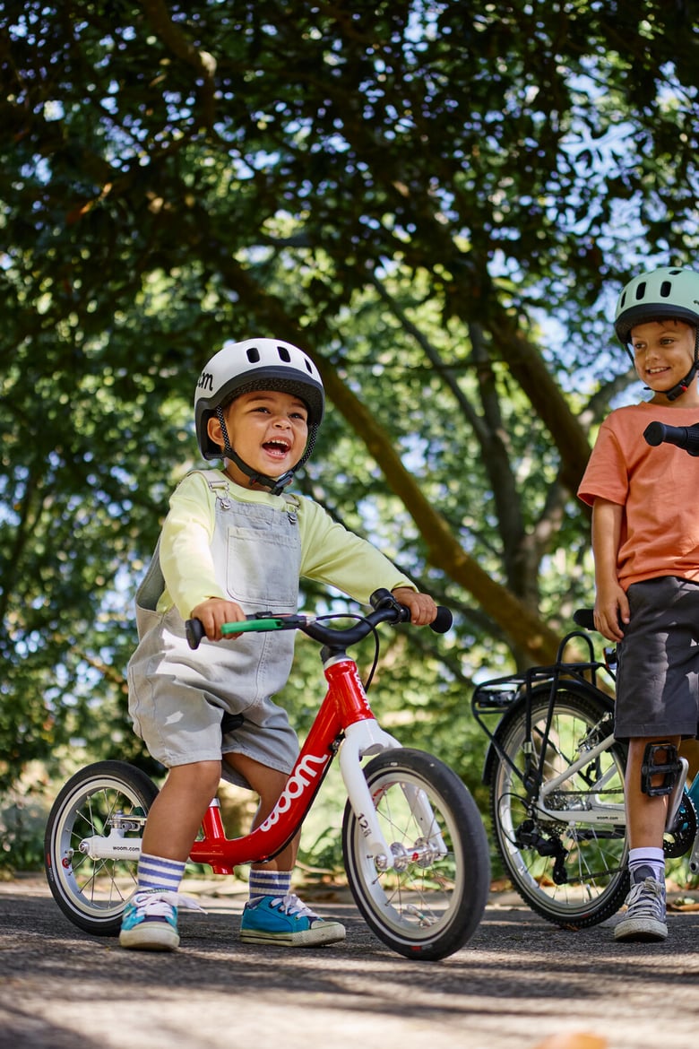 Two smiling children wearing woom READY helmets on their woom GO bikes under trees.