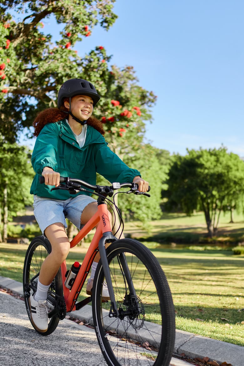 Smiling girl wearing a woom READY helmet on a woom EXPLORE in Neon Coral, riding through a sunny park.