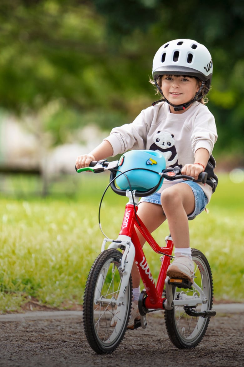 Child in a READY helmet riding a red woom GO bike on a park path.