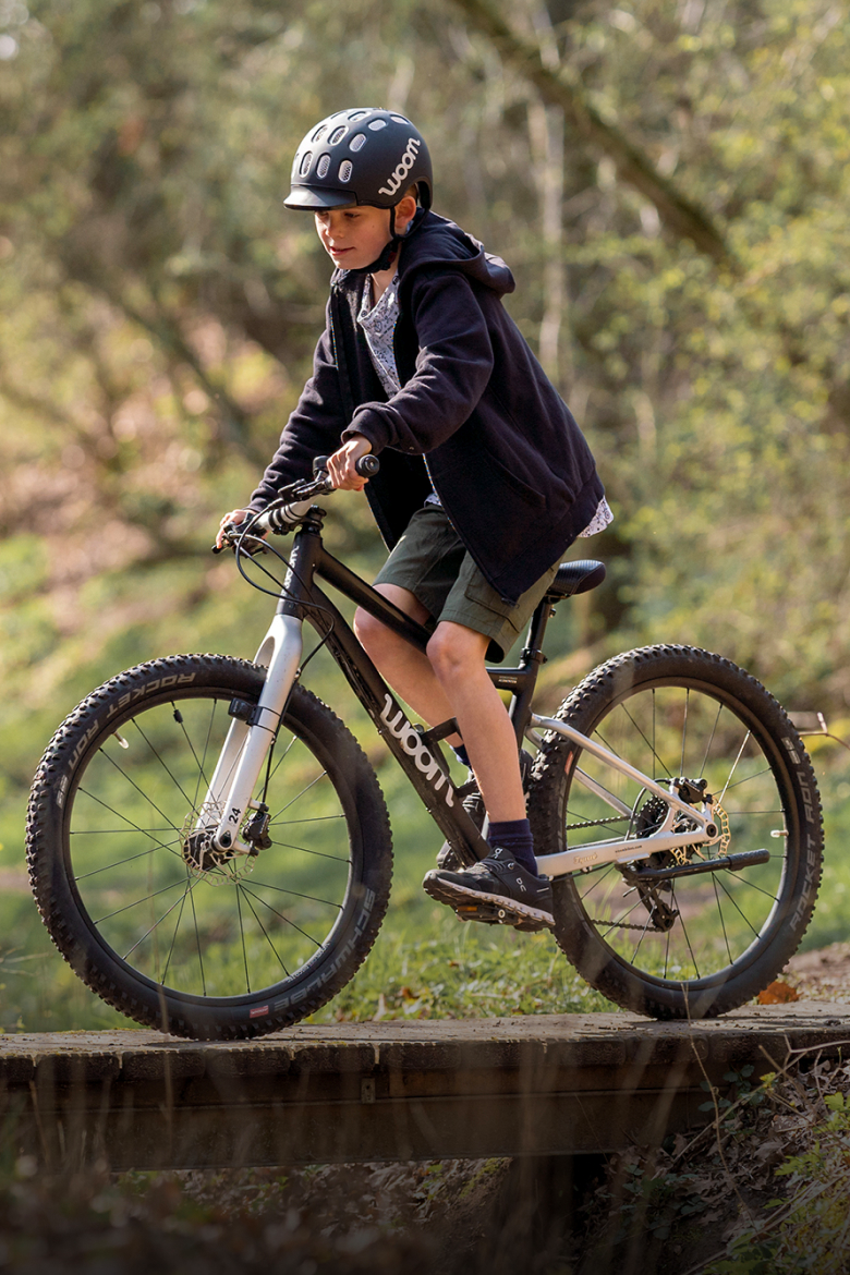 A boy riding a black woom OFF mountain bike wearing a black woom KIDS helmet through a green forest.