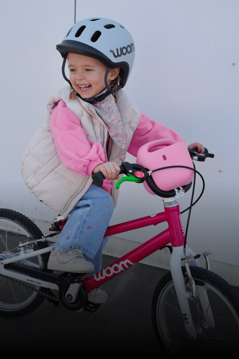 Auf einem rosafarbenen woom GO Kinderfahrrad samt POP Kinderfahrradkorb sitzt ein Mädchen mit grauem READY Helm auf dem Kopf.