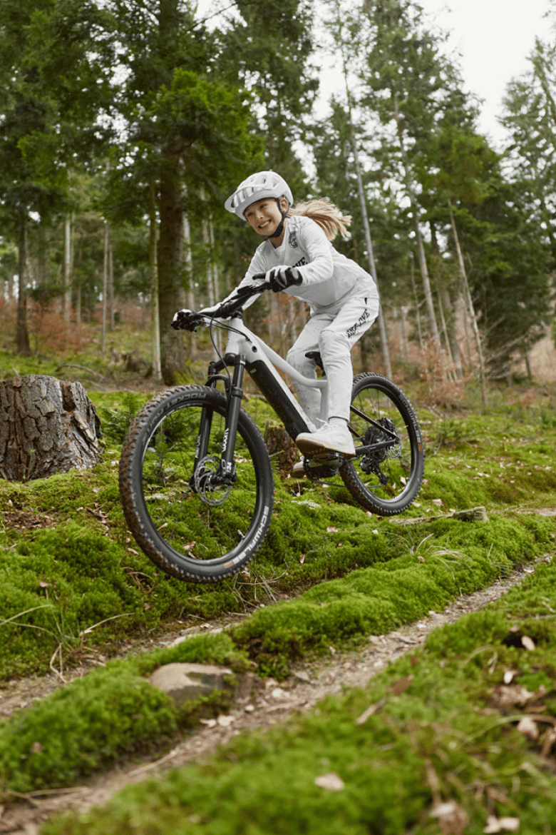 Smiling girl riding the woom UP e-mountain bike in cirrus cloud through a puddle in the forest.