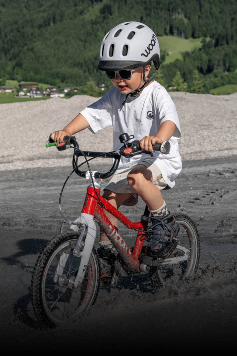 Un enfant avec un casque READY gris et des vêtements d’été traverse un cours d’eau à gué sur son vélo woom rouge.