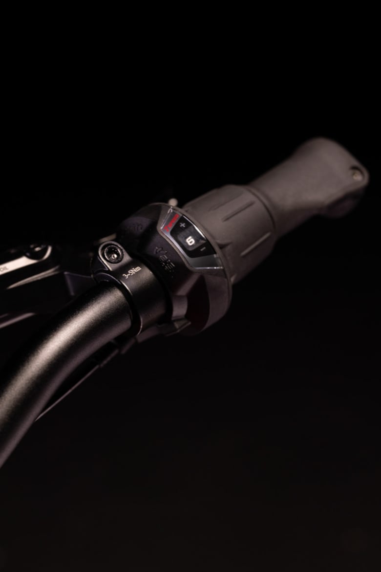 Gros plan du guidon noir woom EXPLORE avec une poignée tournante Shimano noire et un indicateur de vitesse.
